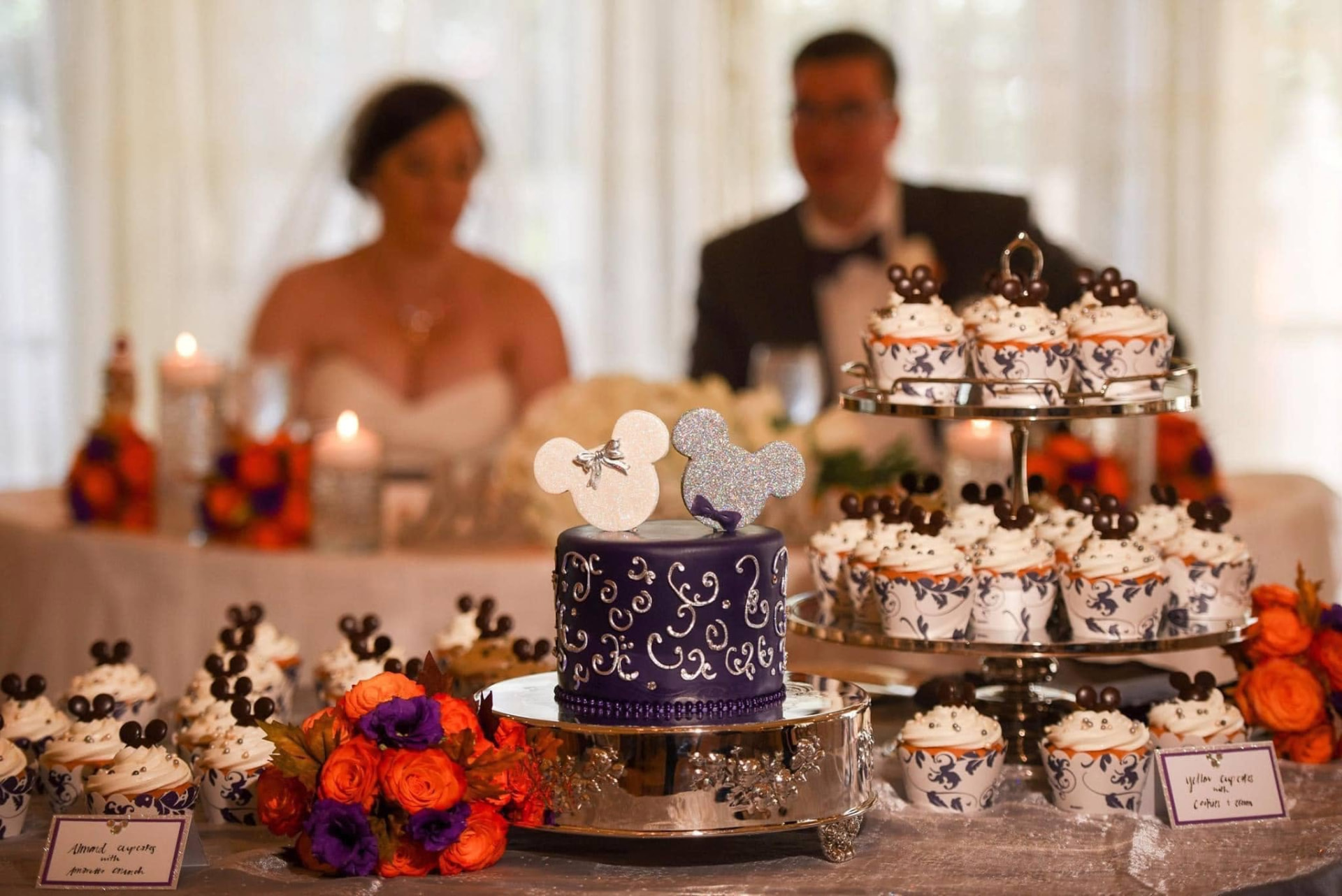Purple Disney wedding cake and Mickey-themed cupcakes with orange and purple floral arrangements